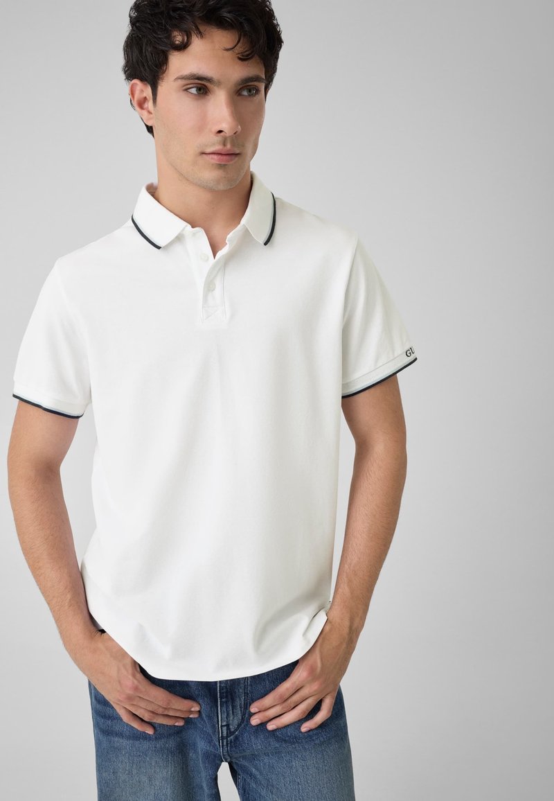 Jeune homme aux cheveux courts et bouclés portant un polo blanc avec des bordures noires et un jean bleu, debout avec les mains près de la taille.