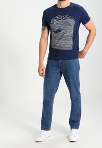 Navy blue t-shirt with a white wave and boat design, paired with light blue jeans and white sneakers; casual fit.