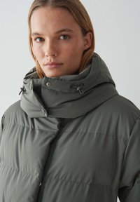 Doudoune en vert atténué, avec un col montant et une capuche ajustable. Texture lisse avec des coutures horizontales à travers le corps.