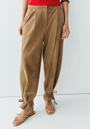 Losse beige broek met enkelbandjes, gedragen met beige teenslippers en een rood topje, afgebeeld van taille tot voeten tegen een effen achtergrond.
