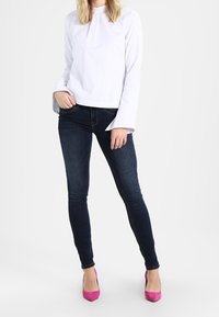 Camiseta blanca de manga larga con puños acampanados, combinada con vaqueros ajustados oscuros y tacones altos de color rosa brillante, sobre un fondo neutro.