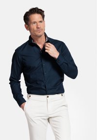 Chemise marine à manches longues avec fermeture boutonn�ée et col intérieur à motifs. Portée avec un pantalon crème ornée d'accents dorés.