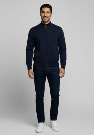 Sudadera navy con cremallera y cuello alto, con puños y dobladillo acanalados; llevada sobre jeans oscuros y zapatillas blancas; textura suave, diseño ajustado.