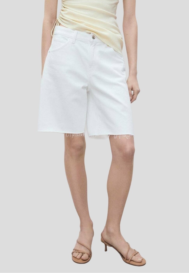 Femme portant un short en denim blanc longueur genou avec des bords à cru et des sandales à talons beiges à brides, debout devant un fond uni.