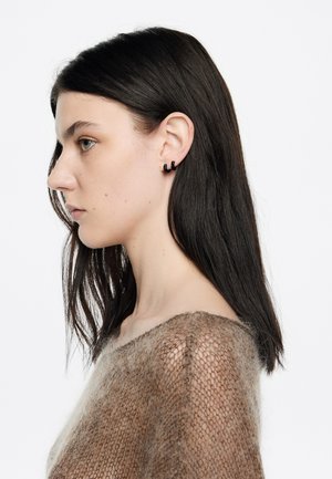 Pendientes circulares negros en una modelo, con un diseño minimalista. Suave suéter marrón de malla resalta la textura y el uso de capas.