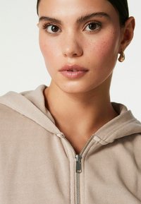 Beige mikina na zip s měkkou texturou, obsahující stříbrný zip a připevněnou kapuci. Detailní pohled na výstřih a pleť s pihami.