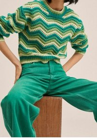 Persoon die een groen, wit en teal chevron-geometrisch gebreide trui en teal wijde broek draagt, zittend op een houten blok met handen op de heupen.