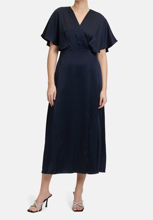 Kobieta ubrana w granatową sukienkę kopertową z rozkloszowanymi rękawami i srebrnymi sandałami na wysokim obcasie, stojąca na tle białego koloru.