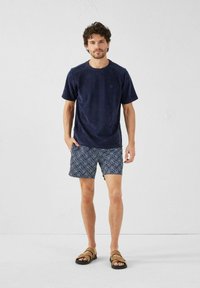 Camiseta de manga corta azul marino de tejido texturizado, combinada con bañadores estampados en azul oscuro y blanco, y sandalias marrones.