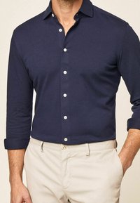 Homme portant une chemise bleu marine à manches longues retroussées et un pantalon beige, une main dans la poche.