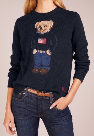 Pull bleu marine avec un ours tricoté portant un jean, des mocassins et un pull noir avec un drapeau américain, associé à un jean bleu et une ceinture marron.