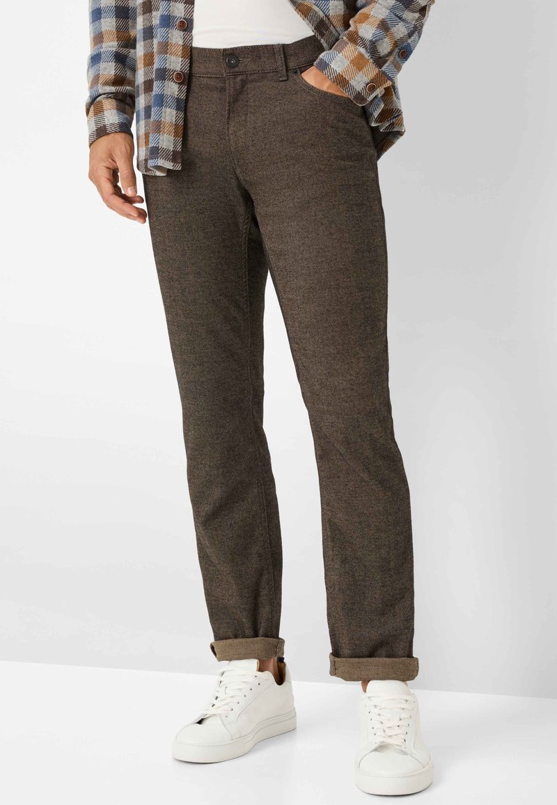 Bruine, op maat gemaakte broek van gestructureerde stof, met zijzakken en een opgerolde zoom. Gecombineerd met witte sneakers voor contrast.