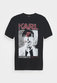 Černé bavlněné tričko s grafikou znázorňující postavu v brýlích, s textem "KARL" a "LEGEND WORLD TOUR" v červené a bílé barvě.