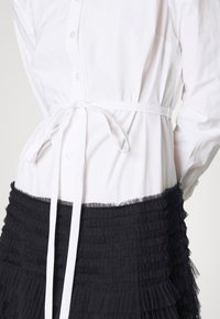 Camicia bianca con colletto abbottonato e vita annodata, abbinata a una gonna in tulle nera a strati con una texture arricciata.