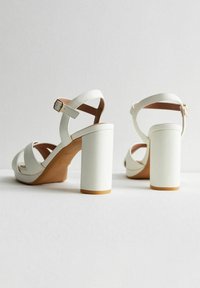 Sandalias blancas de tacón alto con tiras anchas, hebilla en el tobillo y un tacón sólido. Material de cuero suave con plantilla en tono marrón.