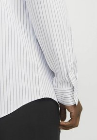 White long-sleeve shirt with navy vertical stripes, featuring a curved hem and buttoned cuffs. Textured fabric with clear stitching details.