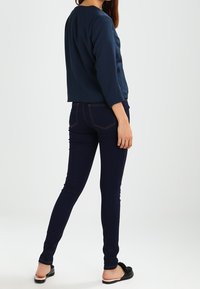 Marineblauwe casual top, lange mouwen, cropped zoom, gecombineerd met donkere skinny jeans en zwarte platte schoenen. Gladde stof met subtiele textuur.