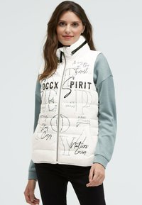 Weiße, gepolsterte Weste mit gesteppten Details, schwarzen Textdrucken und Stehkragen. Die Seiten zeigen ein graues Langarmgarment.