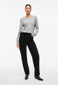 Pilko mezgimo megztinis su plačia iškirpte, derinamas su juodomis plačiomis korduro kelnėmis. Modelis dėvi juodus aulinukus, stovėdamas viena ranka kišenėje.