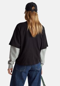Camiseta negra de gran tamaño con mangas grises, que presenta un cuello redondo y un ajuste relajado. Se lleva con jeans de mezclilla azul oscuro y una gorra negra.