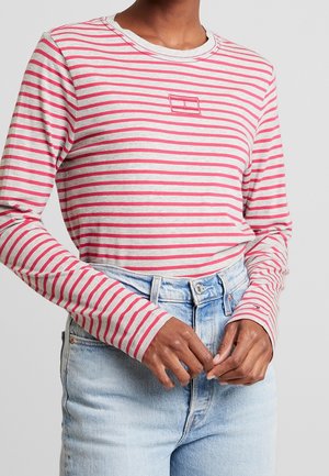 Personne portant une chemise à manches longues rayée rose et blanche rentrée dans un jean taille haute bleu clair sur un fond gris uni.
