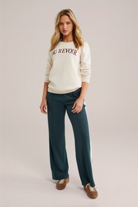 Romige sweatshirt met "AU REVOIR" in bordeauxkleurige letters, gecombineerd met donkergroene wijde broek met witte zijkanten. Bruine sneakers.