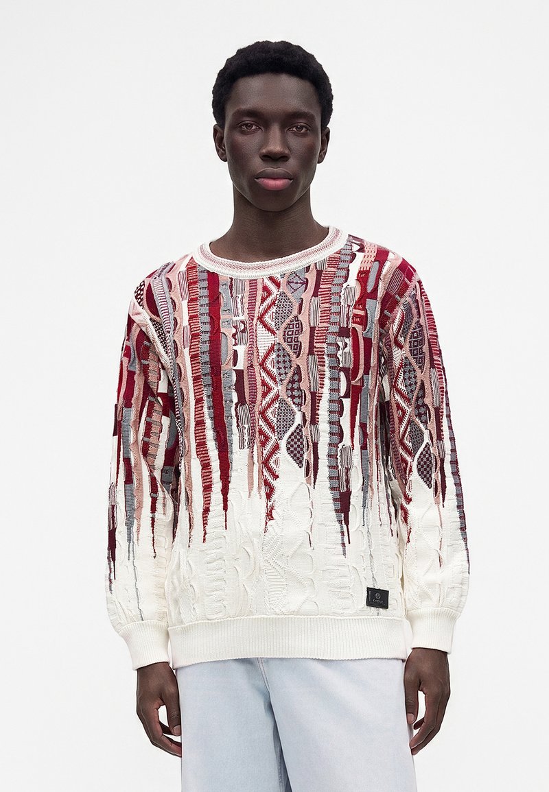Jeune homme portant un pull blanc avec des motifs géométriques verticaux rouges et gris et un pantalon bleu clair, debout devant un fond blanc uni.