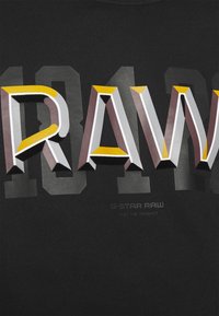 T-shirt nera con testo stampato in grassetto e multicolore "RAW" e numeri, con una texture liscia e grafiche a contrasto.
