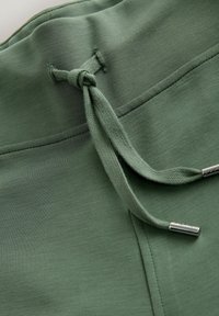 Detalle de cordón verde con terminales metálicos y cintura suave, que presenta una textura de tela ligera y suave, y un diseño sencillo.