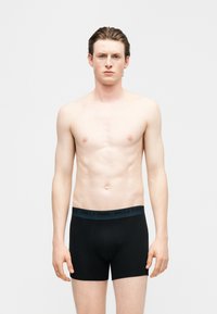 Νέος άνδρας με ανοιχτόχρωμο δέρμα και μαλλιά χτενισμένα προς τα πίσω στέκεται γυμνόστηθος φορώντας μαύρα εσώρουχα boxer με ζώνη μέσης "Only & Sons" σε απλό φόντο.