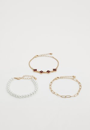 Trois bracelets : chaîne en or avec des pierres précieuses rouges, chaîne en or avec maillons, et perles blanches avec fermoirs en or sur fond blanc.