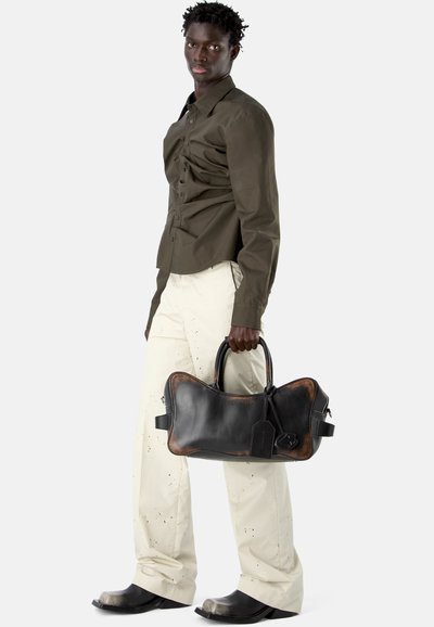 Homme portant une chemise vert olive et un pantalon beige clair tenant un sac en cuir noir et marron en forme d'os.