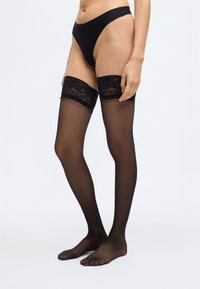 Czarne pończochy do uda z przezroczystym wykończeniem, koronkowym pasem na górze i elastycznym obszyciem. Charakteryzują się gładką fakturą i odkrytymi palcami.