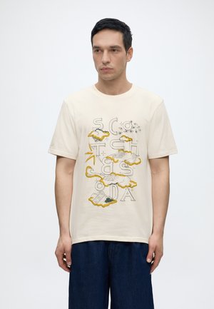 Homme debout portant un t-shirt crème avec des motifs abstraits de nuages jaunes et noirs ainsi que des grandes lettres contours éparpillées, associé à un pantalon bleu foncé.