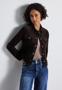 Veste en velours côtelé marron foncé avec des boutons en métal, deux poches poitrine et un col, associée à un haut beige clair et un jean bleu.