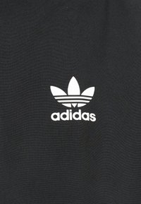 Λευκό λογότυπο Adidas trefoil με τρεις οριζόντιες ρίγες και κείμενο με πεζά γράμματα σε μαύρο ύφασμα.
