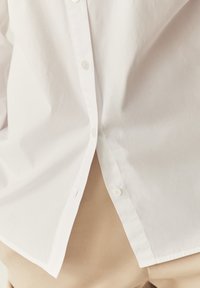Chemise blanche boutonnée en coton. Présente de petits boutons, une texture lisse et un ourlet arrondi. Associée à un pantalon beige en bas.