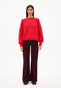 Rot gestrickter Pullover mit strukturiertem Muster und gerippten Bündchen, kombiniert mit dunkelburgunderfarbenen Cordhosen mit weitem Bein. Schwarze Schuhe runden den Look ab.
