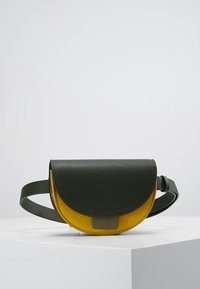 Mała torebka na pasek w kształcie półksiężyca z ciemnozieloną skórzaną klapką i paskiem, musztardowożółtym zamszowym korpusem, prezentowana na białym piedestale.