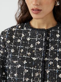 Giacca di tweed nera con dettagli floreali metallici, cuciture bianche a contrasto, bottoni rotondi e un collo sfrangiato.