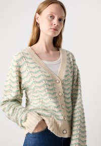 Cardigã texturizado em creme com listras onduladas em turquesa, apresentando um decote em V, botões semelhantes a pérolas e punhos e bainha canelados.