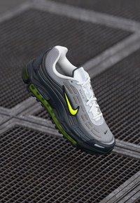 Nike Air Max kurpes ar gaisa plīslu gaiši pelēkā krāsā, ar melniem akcentiem un spilgti dzeltenu logo. Bieza amortizācija ar zaļām zoles detaļām.