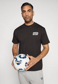 T-shirt preta com "JUVENTUS MILLE897" em branco, combinada com calças de moletom cinzentas; segurando uma bola de futebol branca com realces em azul e preto.