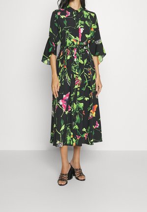 Mujer con un vestido midi negro con estampado colorido de mariposas y flores, cintura con cinturón y sandalias negras de tacón con tiras cruzadas.