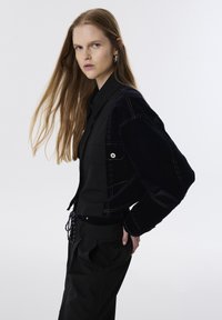 Zwarte fluwelen jas met contrasterende witte stiksels, zijzakdetail, gecombineerd met een zwart shirt en losse broek.