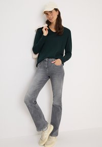 Grünes Longsleeve-Oberteil mit V-Ausschnitt, graue Jeans und hellfarbene Sneakers. Trägt eine cremefarbene Mütze und eine beigen Oberbekleidung, die über die Schulter gelegt ist.