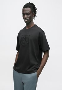 Μαύρο βαμβακερό T-shirt με κοντά μανίκια, με ανάγλυφο λογότυπο Calvin Klein στο κέντρο, συνδυασμένο με ανοιχτό γκρι αθλητικό παντελόνι.