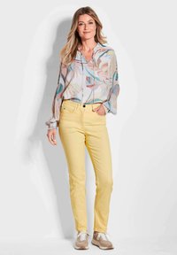 Leichte, gemusterte Bluse mit Blätterdesign in Pastellfarben; kombiniert mit gelben, slim-fit Hosen; braune Sneakers runden das Outfit ab.