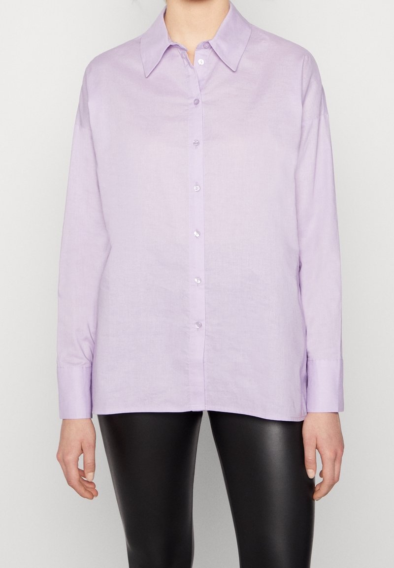 Chemise boutonnée violet clair avec col pointu, manches longues et texture lisse, associée à des leggings noirs. Design simple, sans motifs.