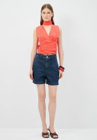 Top rojo sin mangas con un escote en V profundo y textura fruncida, combinado con pantalones cortos de mezclilla oscuros de cintura alta y sandalias marrones de dedo.